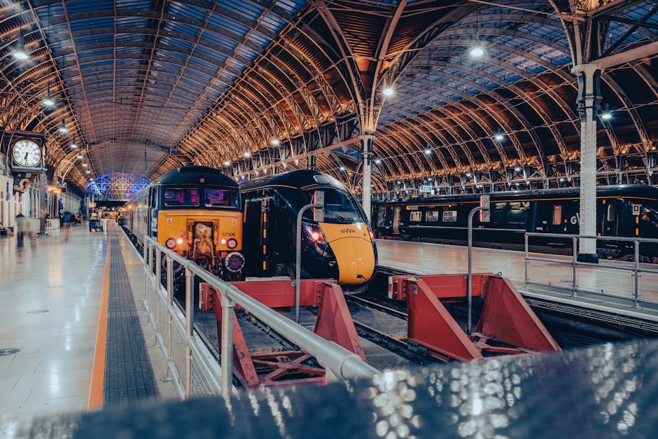 Inside a large train station with a curved glass and metal ceiling allowing natural light to illuminate the area. The platform features various white support columns and a yellow and black train is ap