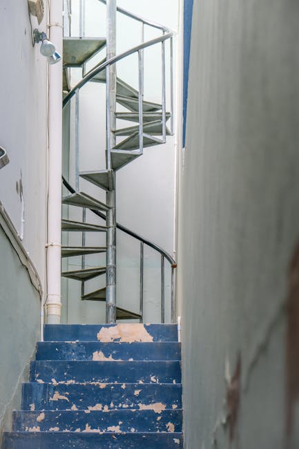 A narrow outdoor staircase made of stone steps leading upward between two walls, with metal handrails attached on both sides. On the left side, the wall is lined with corrugated metal sheeting, while
