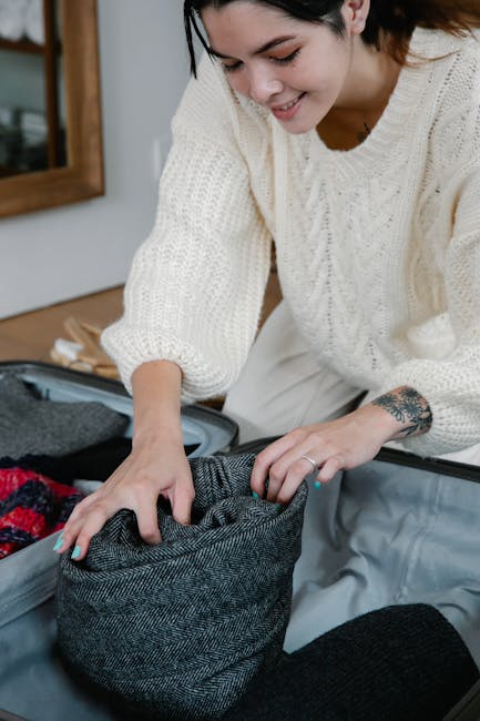 A woman with short dark hair and a tattoo on her wrist is smiling while packing a grey fabric bag with clothing or personal items, inside a home environment with wooden furniture and a mirror in the background. She is wearing a cream-colored knitted sweater and is bent over a large open suitcase on the floor, preparing for a house move or home relocation. The scene depicts packing and moving preparations, with other packing materials and clothes visible nearby, highlighting the process of organizing belongings for a house removal or professional moving service such as Paddington Movers.