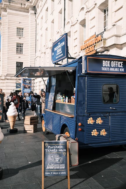 A blue mobile food and ticket sales kiosk is parked on a busy street in central London, adjacent to a white stone building with tall windows. The kiosk has a small service window with a wooden countertop, where various bottles and containers are arranged. A blackboard sign in front of the kiosk displays ticket purchase information, indicating it is open seven days a week. In the background, several pedestrians walk past, some carrying shopping bags or backpacks, and a large ice cream cone sculpture is visible on the sidewalk. Cardboard boxes and wooden crates are placed on the pavement near the kiosk, suggesting ongoing moving or delivery activity. The scene is well-lit with natural daylight, capturing a typical outdoor setting suitable for house removals and furniture transport, as managed by Paddington Movers, focusing on efficient packing and loading processes near Praed Street, Paddington.